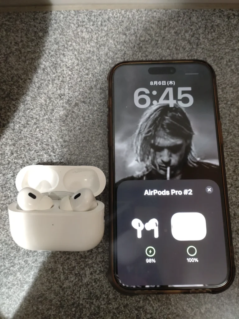 「iPhone 15 Pro Maxの画面にAirPods Proのバッテリー残量ポップアップが表示され、横にケースに入ったAirPods Proが置かれている様子」