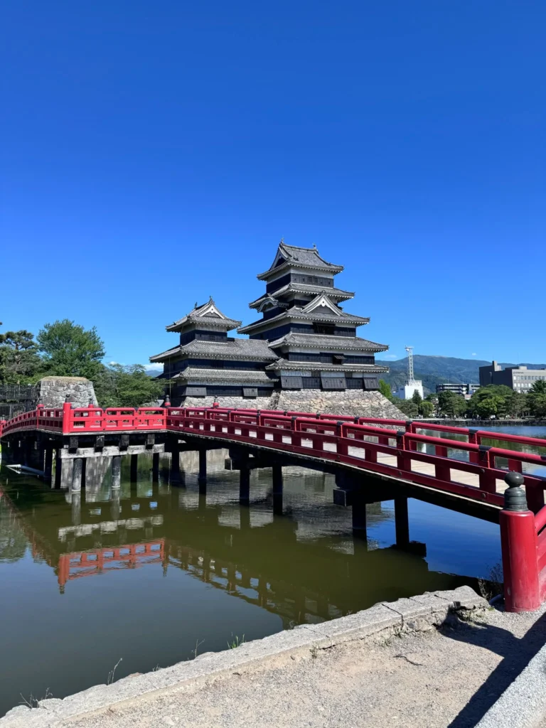 澄み切った青空の下、水堀に架かる赤い橋と黒い天守閣がそびえ立つ松本城